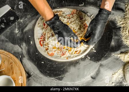 Cooking pizza on a steel table; dough, flour, pizza topping, sauce and chef`s hands in black gloves on the table Stock Photo